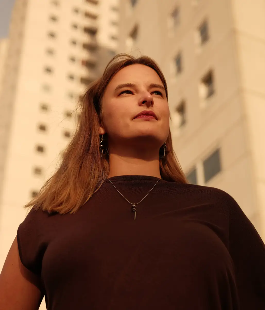 Een portret van een vrouw, ze is van onderaf gefotografeerd en kijkt in de verte. Achter haar zien we hoge industriele gebouwen.