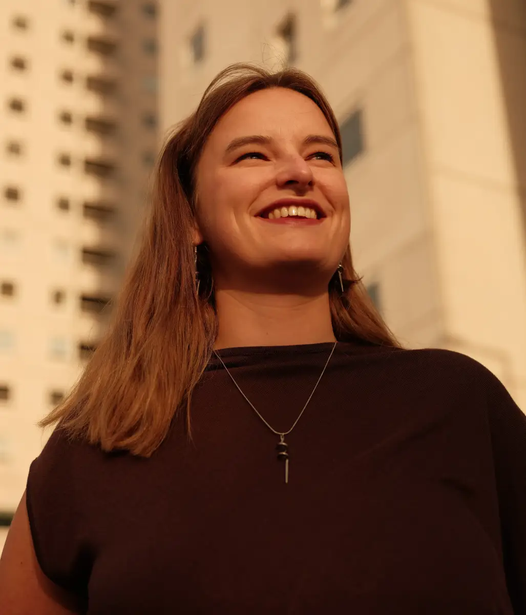 Een portret van een vrouw, ze is van onderaf gefotografeerd en ze kijkt lachend in de verte. Achter haar zien we hoge industriele gebouwen.