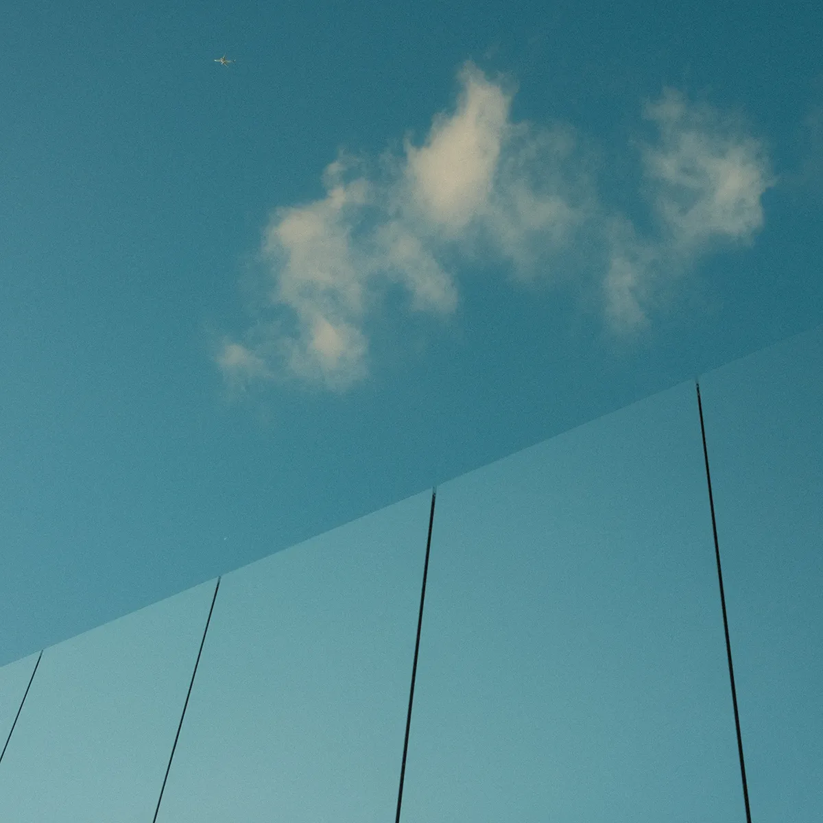 Een foto van een weerspiegeld gebouw, we zien enkel een weerspiegeling van een strak blauwe lucht, het is bijna niet meer duidelijk waar de reflectie stopt en de echte lucht begint. We zien een eenzame wolk boven het gebouw langs zweven.