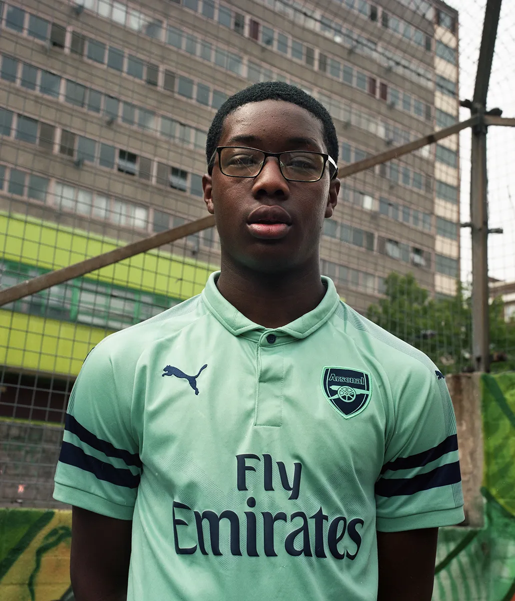 Een portret van een jongen in een voetbalshirt, hij staat in een voetbalcourt en kijkt recht in de camera, achter hem zien we een enorm flatgebouw waar hij woont in Londen.