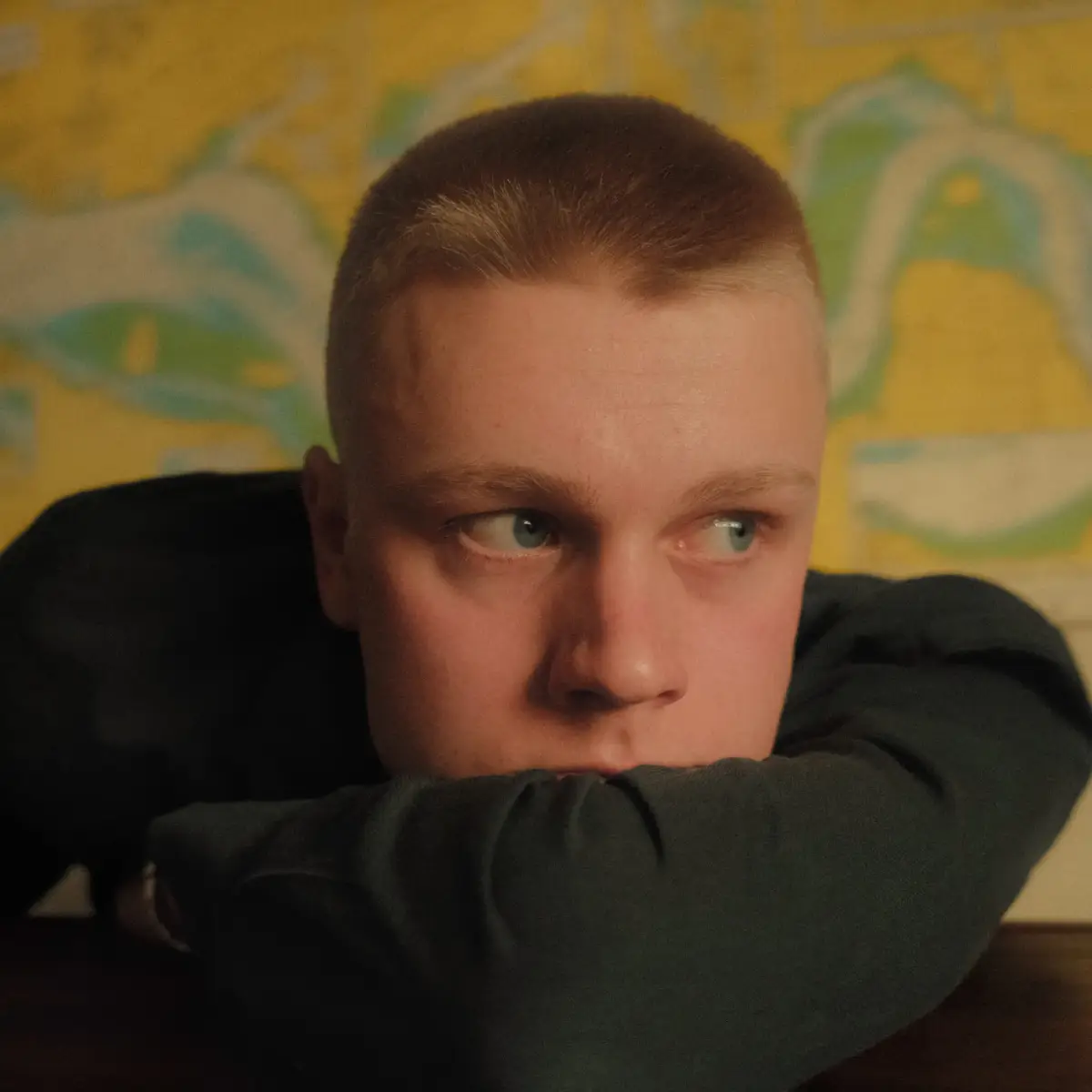 Close-up portret van een jongen man die op zijn arm leunt en rechts het beeld uit kijkt. Hij heeft een geschoren hoofd.