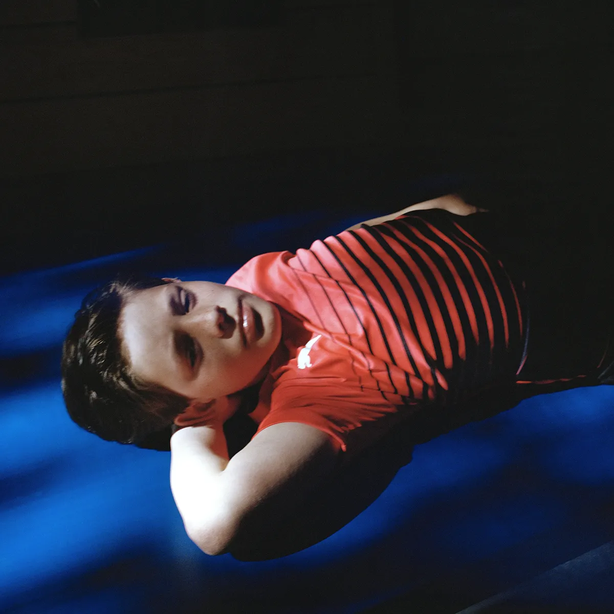 Een portret van een jonge jongen die in een gymzaal op de grond ligt, hij ligt op een arm en het licht van door een raam perfect op zijn gezicht, hij kijkt dromerig de ruimte in.