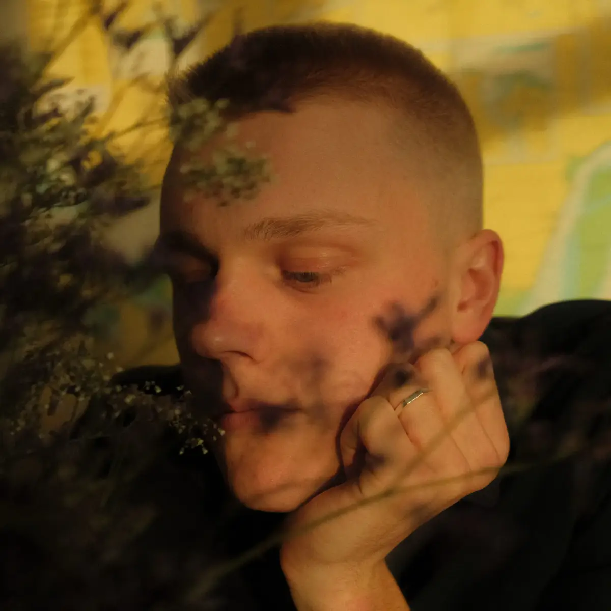 Close-up portret van een jongen man die op zijn arm leunt en linksonder het beeld uit kijkt. Voor hem zien we wat bloemen die een deel van het beeld bedekken, we zien hem door de bloemen heen. Hij heeft een geschoren hoofd.