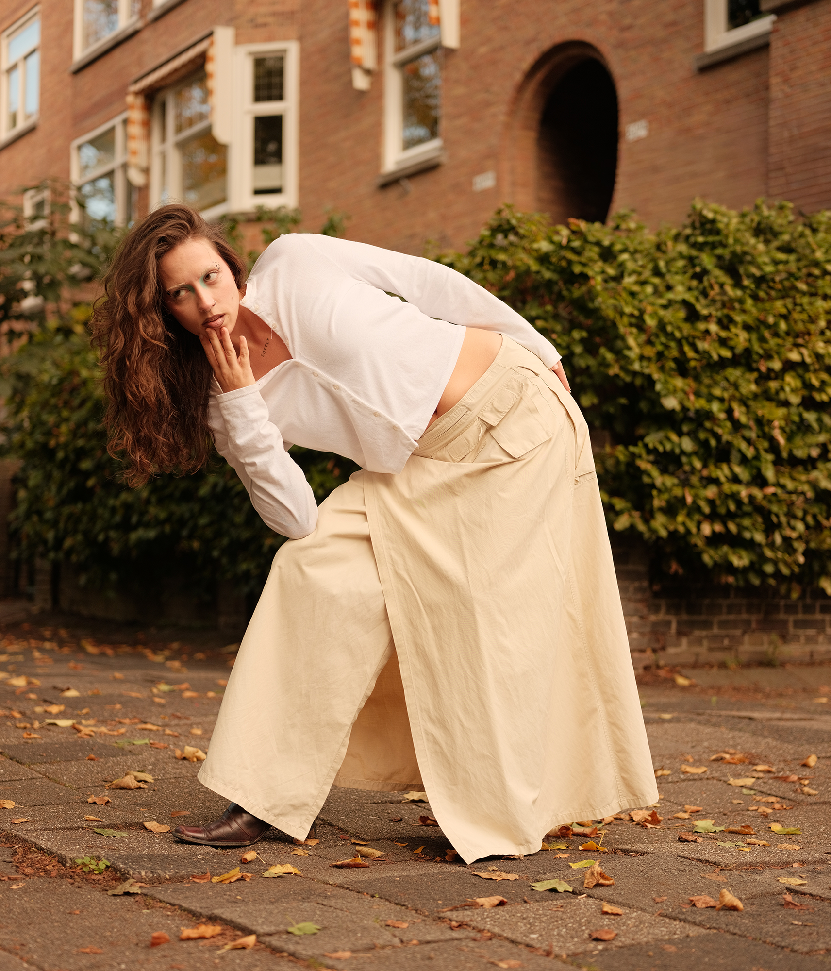 Een portret van een dame met uitgesproken make-up, in een prachtige outfit, ze staat uniek geposeert, met haar arm hangt ze op haar gebogen knie voorover. Ze kijkt recht in de camera.
