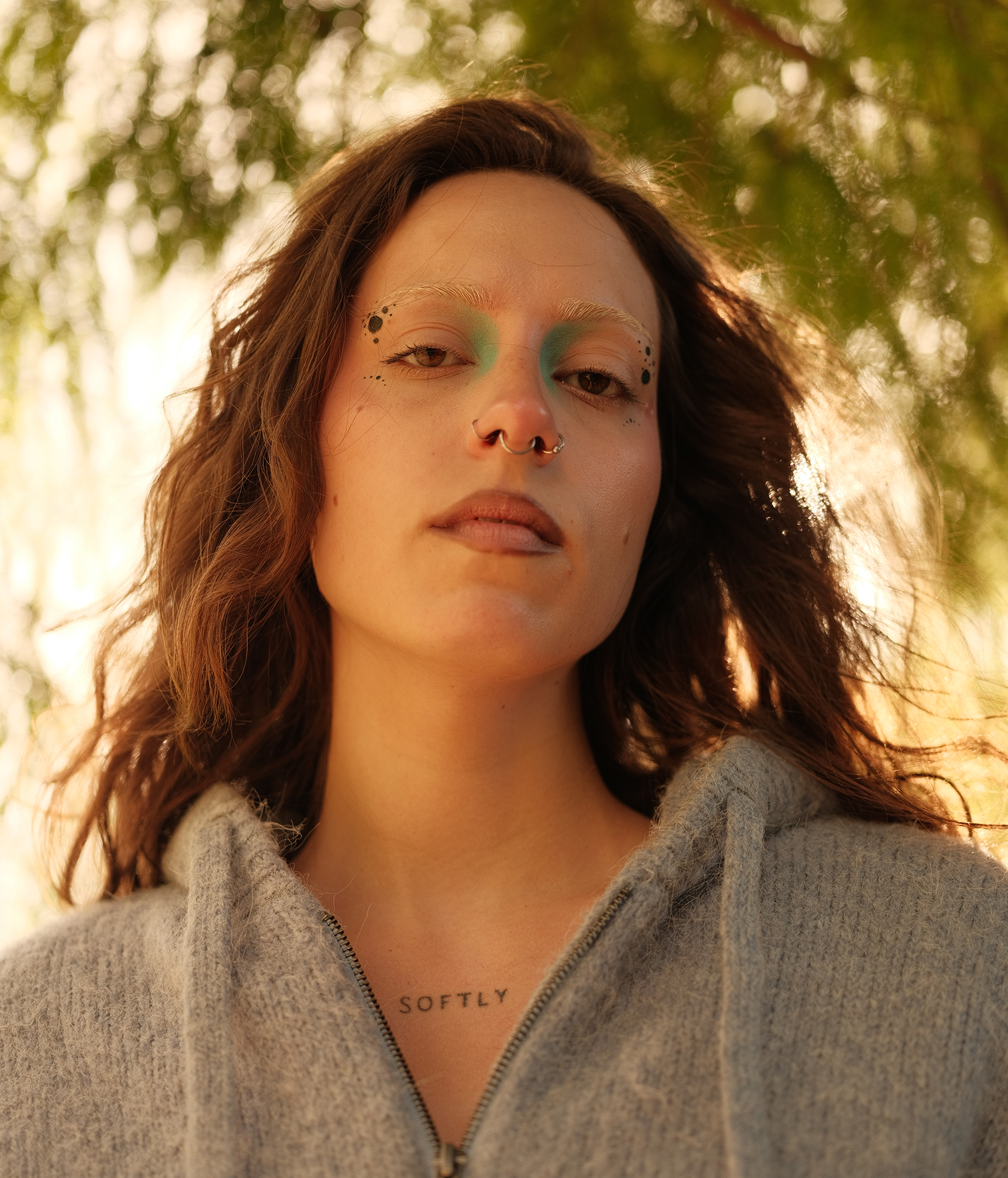 Een portret van een dame met uitgesproken make-up, in een mooi park zien we achter haar hoofd het licht door de bomen komen, het perspectief is vanaf onder