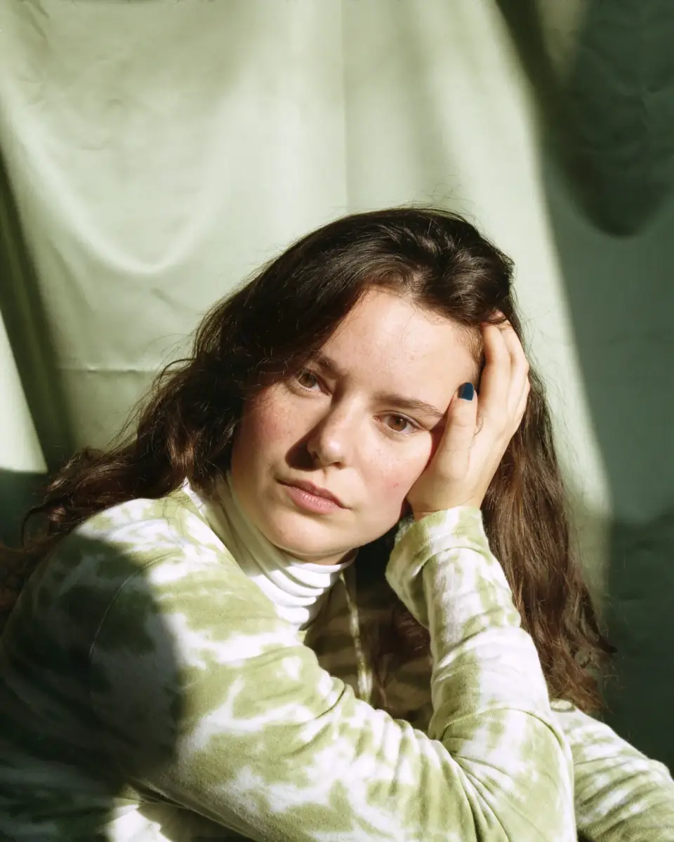 Portret van een jonge dame die in een groen shirt voor een groene achtergrond zit, ze leunt op haar hand en kijkt in de camera