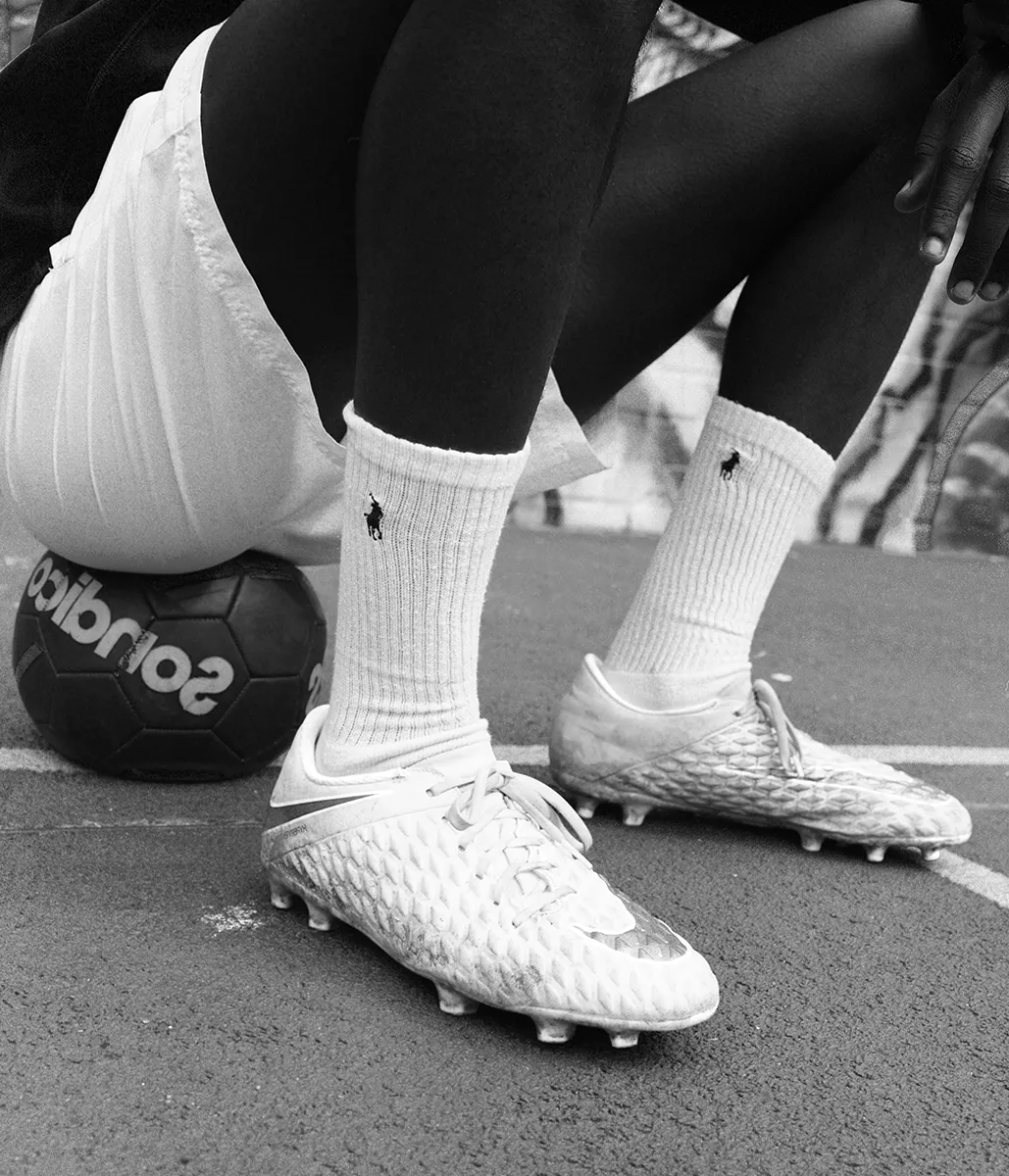 Een zwart-wit foto van een jongen die op een voetbal zit, we zien zijn benen en zijn sportschoenen nadrukkelijk in beeld.