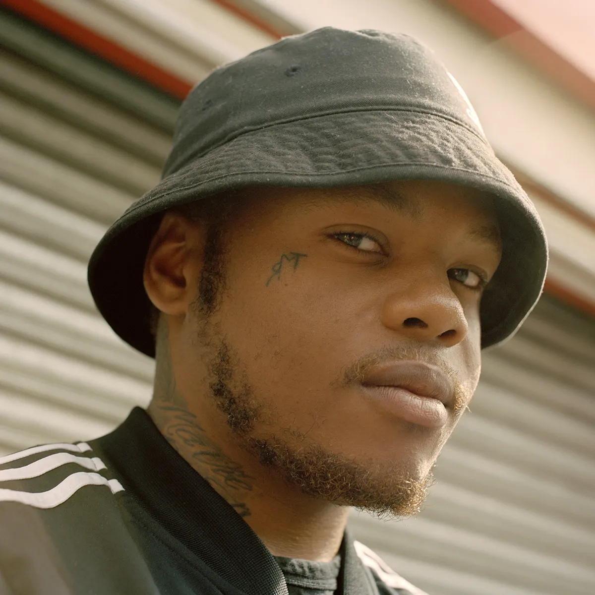 Een portret van een jonge man met gezichtstatoeages, hij kijkt recht in de camera en heeft een zwarte bucket hat op.